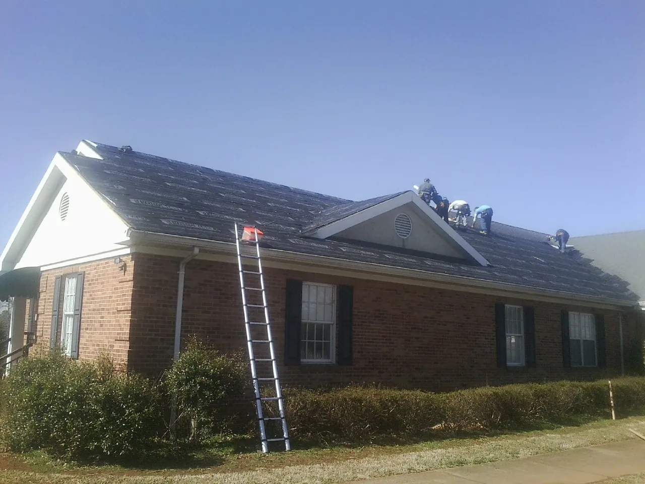 Crew tearing shingles off a brick ranch roof with a ladder set — Metal Roof Repair work by our crew in Kohler