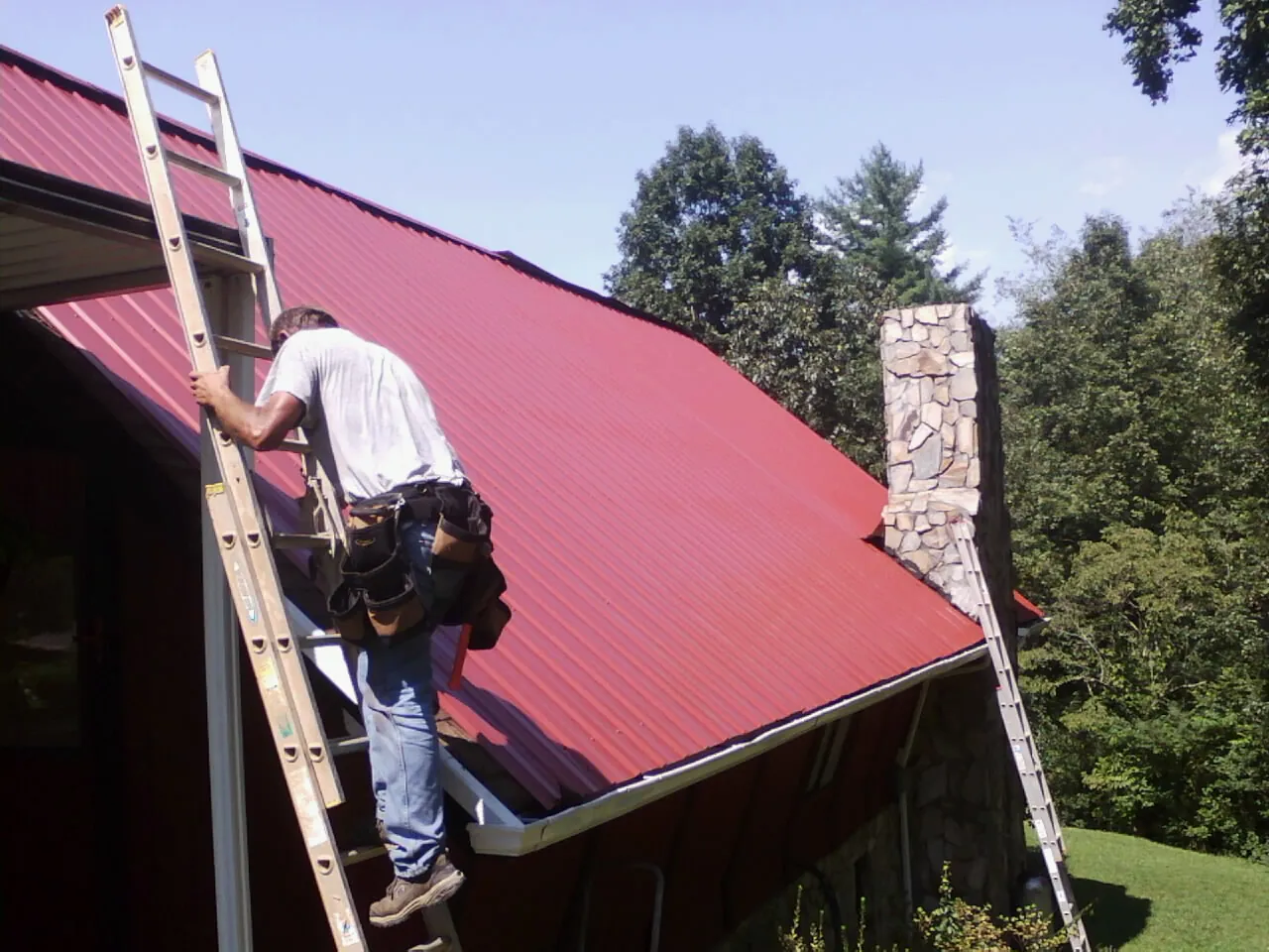 Roofer on a ladder beside a red metal roof and stone chimney — Soffit & Fascia Repair by Roofing Brothers in Kohler
